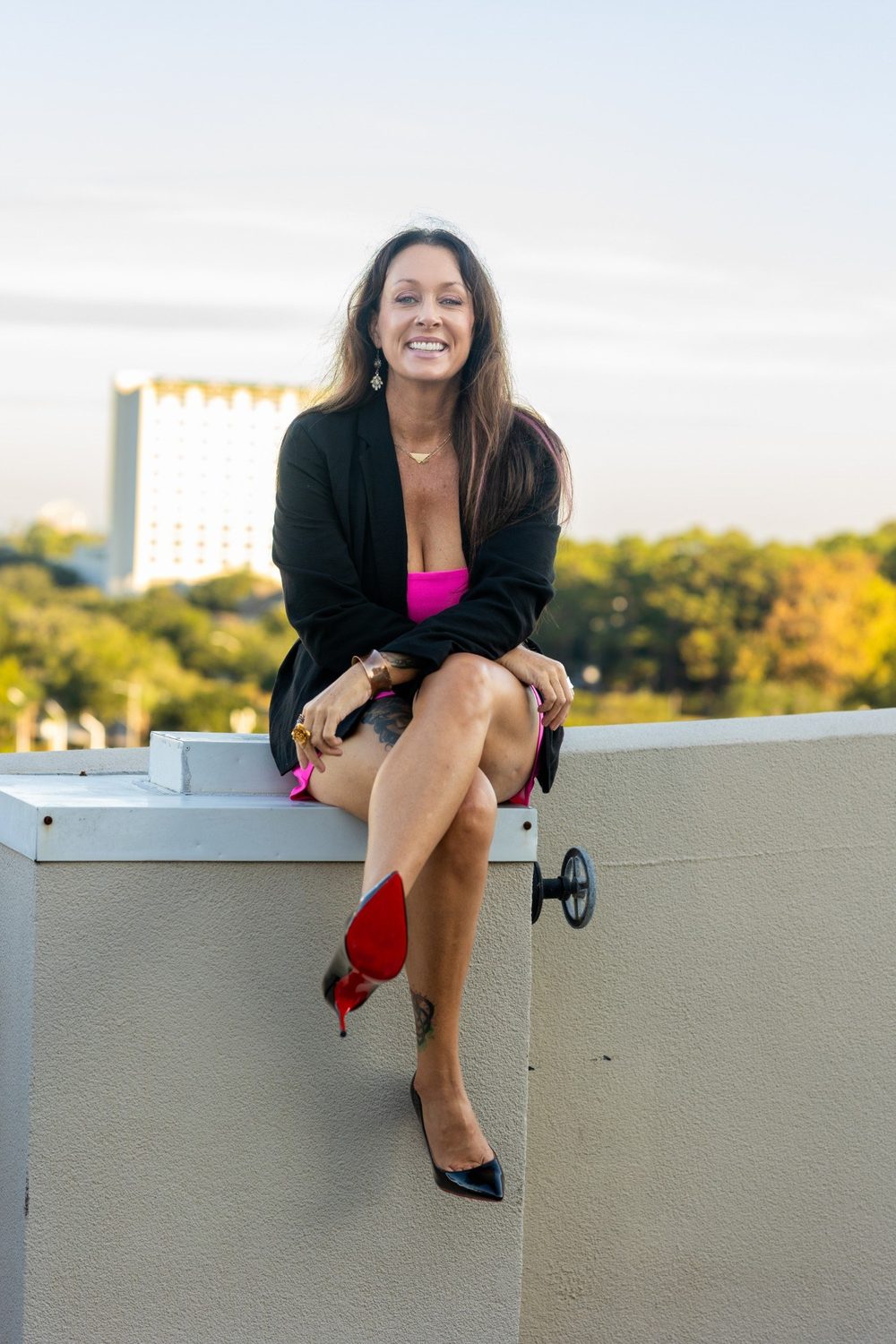 Leslie M Lyon, fractional CMO and founder of Leslie M Lyon, sitting on a rooftop ledge at sunset in a black blazer and pink dress